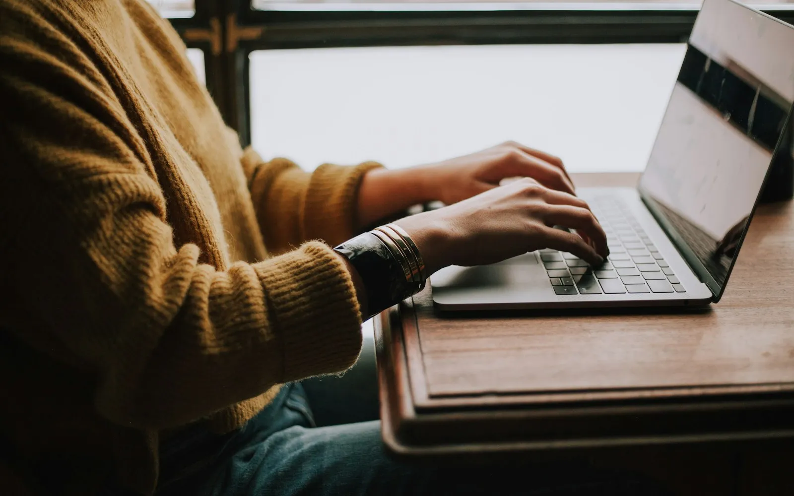 Person in a mustard sweater typing on a laptop at a wooden table by a window